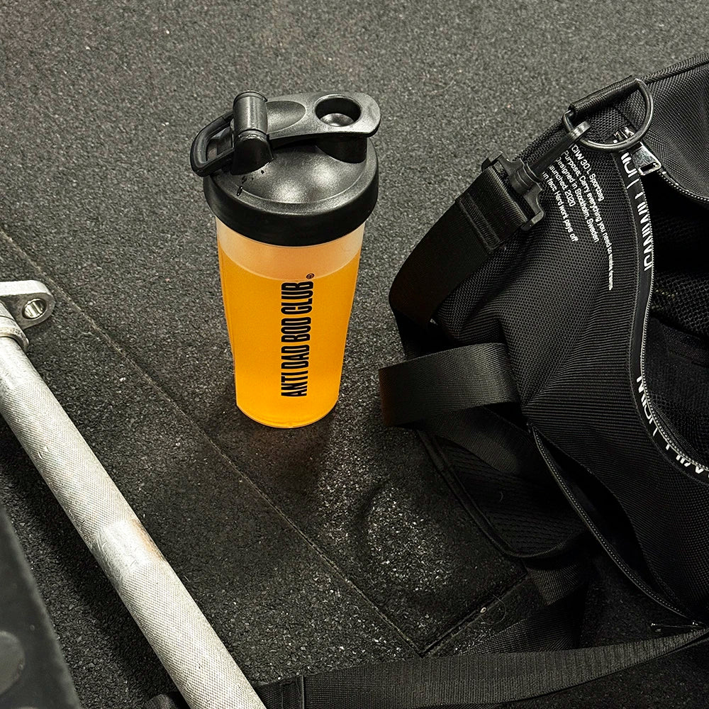 Orange shaker bottle with black cap on a textured surface next to a black bag.