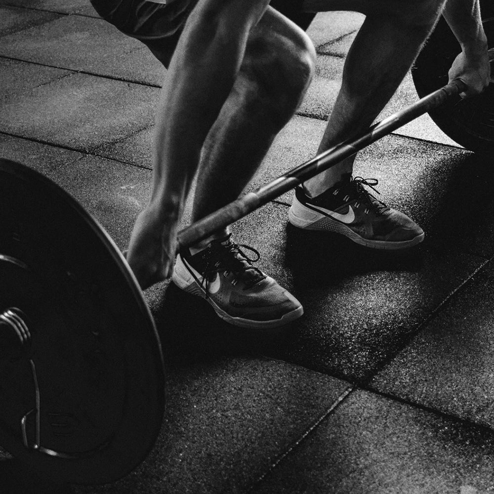 Person lifting a barbell with focus on feet and shoes on a tiled floor.