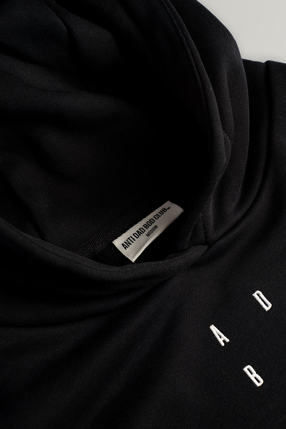 Close-up of a black hooded sweatshirt with a white interior neck label reading 'ANTI DAD BOD CLUB' and white cropped letters on the chest.