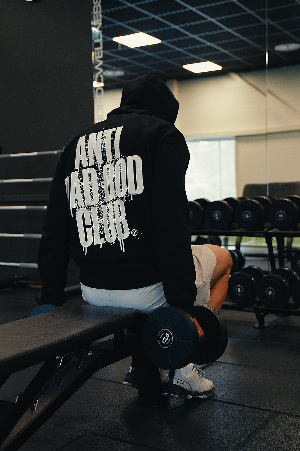 A person in a black hoodie sits on a gym bench holding a dumbbell, showing a large white graphic on the back that reads 'ANTI DAD BOD CLUB'. The gym has weight racks and mirrors in the background.