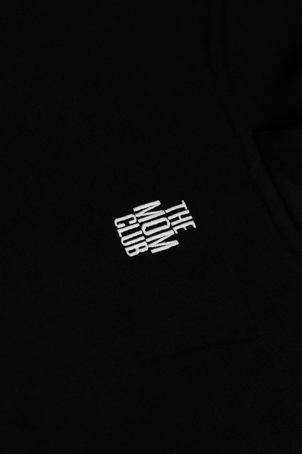 Close-up of a black garment featuring a white embroidered graphic on the chest that reads 'THE MOM CLUB' in stacked, stylized lettering.